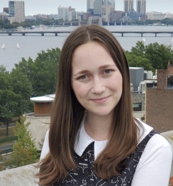 A color photograph of a person's head and shoulders. The person is facing the camera and looking directly at the viewer. They have long brown hair and are wearing a black dress with white constellations. The background is of a river with skyscrapers and other buildings behind it. The image is a portrait-style headshot, commonly used for professional purposes such as resumes, online profiles, or business cards.